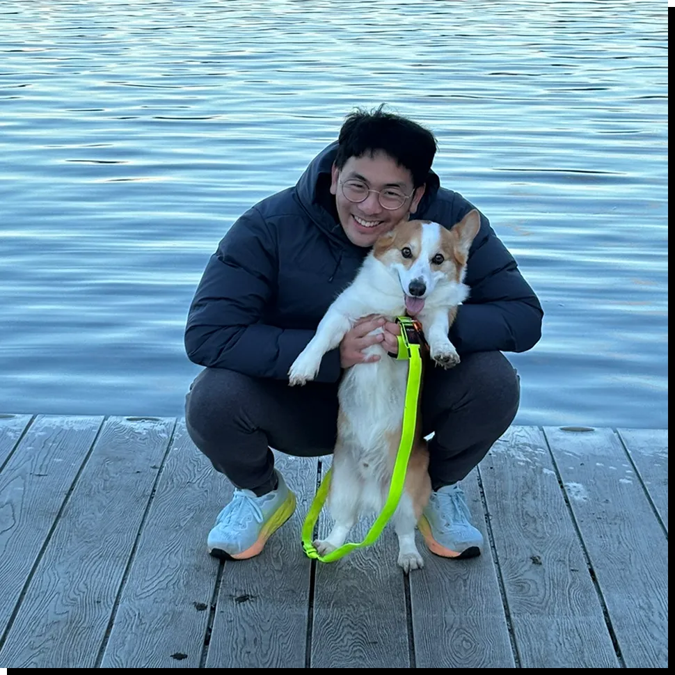 Darren crouching on a dock by the water, holding a corgi